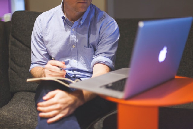 Man sits at an Apple computer with notebook and legs crossed